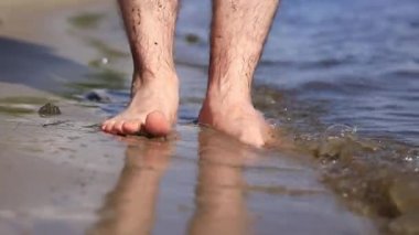 Sahilde çıplak erkek ayakları. Yazın nehrin yanında yürü. Suya yakın kumda duran insan ayaklarının yakın çekimi. Kumsal. Yazın nehirde dinlen. Adam ayaklarını nehir suyunda ıslatıyor.