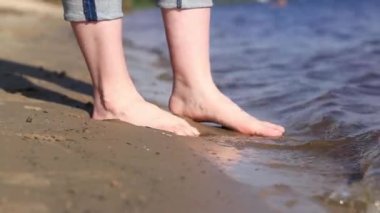 Sahilde çıplak kadın ayakları. Yazın nehrin yanında yürü. Suya yakın kumda duran insan ayaklarının yakın çekimi. Kumsal. Yazın nehirde dinlen. Bir kadın ayaklarını nehir suyunda ıslatıyor.
