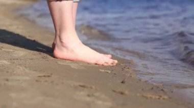 Sahilde çıplak kadın ayakları. Yazın nehrin yanında yürü. Suya yakın kumda duran insan ayaklarının yakın çekimi. Kumsal. Yazın nehirde dinlen. Kadın ayaklarını nehir suyunda ıslatıyor.