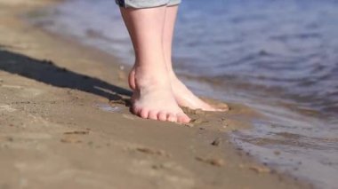 Sahilde çıplak kadın ayakları. Yazın nehrin yanında yürü. Suya yakın kumda duran insan ayaklarının yakın çekimi. Kumsal. Yazın nehirde dinlen. Kadın ayaklarını nehir suyunda ıslatıyor.