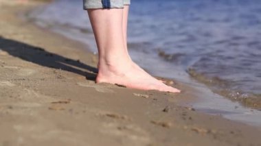 Sahilde çıplak kadın ayakları. Yazın nehrin yanında yürü. Suya yakın kumda duran insan ayaklarının yakın çekimi. Kumsal. Yazın nehirde dinlen. Bir kadın ayaklarını nehir suyunda ıslatıyor.