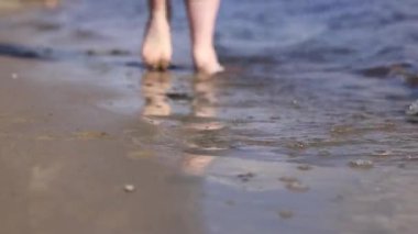 Sahilde çıplak kadın ayakları. Yazın nehrin yanında yürü. Suya yakın kumda duran insan ayaklarının yakın çekimi. Kumsal. Yazın nehirde dinlen. Bir kadın ayaklarını nehir suyunda ıslatıyor.