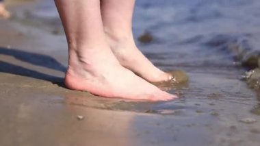 Sahilde çıplak kadın ayakları. Yazın nehrin yanında yürü. Suya yakın kumda duran insan ayaklarının yakın çekimi. Kumsal. Yazın nehirde dinlen. Bir kadın ayaklarını nehir suyunda ıslatıyor.