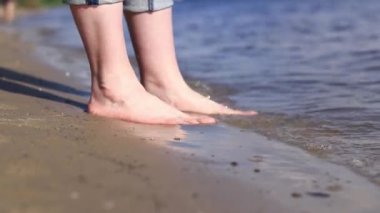 Sahilde çıplak kadın ayakları. Yazın nehrin yanında yürü. Suya yakın kumda duran insan ayaklarının yakın çekimi. Kumsal. Yazın nehirde dinlen. Bir kadın ayaklarını nehir suyunda ıslatıyor.