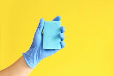 Bulaşık süngerli mavi lateks eldivenli bir adam eli. Mavi sünger elinde, odanın ıslak temizliği, sarı arka plan. Temizlik. Bulaşıkları yıka. Temizlik servisi, temizlik.