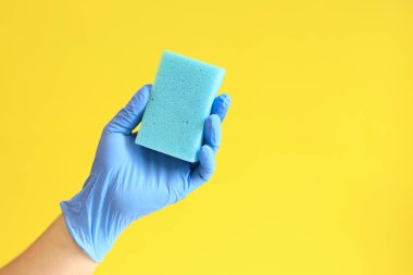 Bulaşık süngerli mavi lateks eldivenli bir adam eli. Mavi sünger elinde, odanın ıslak temizliği, sarı arka plan. Temizlik. Bulaşıkları yıka. Temizlik servisi, temizlik.
