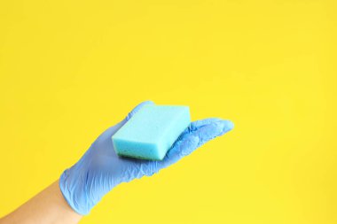 Bulaşık süngerli mavi lateks eldivenli bir adam eli. Mavi sünger elinde, odanın ıslak temizliği, sarı arka plan. Temizlik. Bulaşıkları yıka. Temizlik servisi, temizlik.