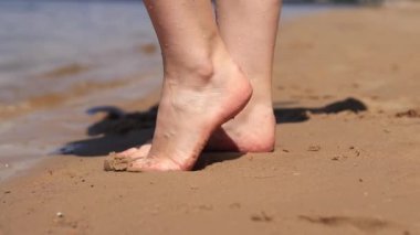 Sahilde çıplak kadın ayakları. Yazın nehrin yanında yürü. Suya yakın kumda duran insan ayaklarının yakın çekimi. Kumsal. Yazın nehirde dinlen. Bir kadın ayaklarını nehir suyunda ıslatıyor.