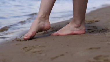 Sahilde çıplak kadın ayakları. Yazın nehrin yanında yürü. Suya yakın kumda duran insan ayaklarının yakın çekimi. Kumsal. Yazın nehirde dinlen. Bir kadın ayaklarını nehir suyunda ıslatıyor.