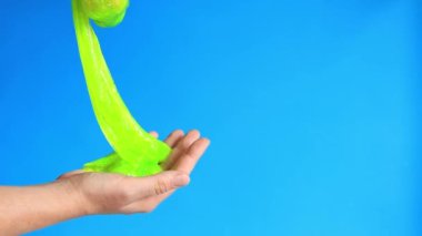 Kadının elleri mavi arka planda parlak yeşil bir balçığı birbirine yaklaştırıyor. Rahatlamak için stres önleyici oyuncak. Eğlenceli duyusal aktivite. Oyuncakçı konsepti. Kız yeşil balçık yoğuruyor.