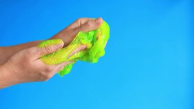 Kadının elleri mavi arka planda parlak yeşil bir balçığı birbirine yaklaştırıyor. Rahatlamak için stres önleyici oyuncak. Eğlenceli duyusal aktivite. Oyuncakçı konsepti. Kız yeşil balçık yoğuruyor.