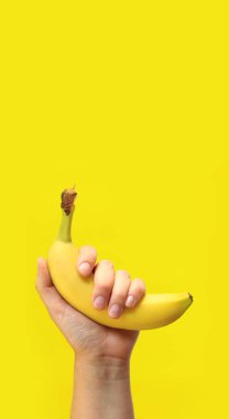 Sarı arka planda bir kadının elinde muz. Olgunlaşmış muzlu bir elin yakın çekimi. Sağlıklı beslenme, vitamin ve diyet kavramı. Meyveler. Beyaz kadın eli kabuktaki muzu gösteriyor.