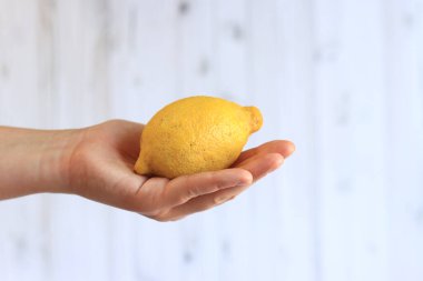 Hafif ahşap arka planda bir kadının elinde olgun bir limon. Limonlu bir elin yakın çekimi. Hasat et. Sağlıklı beslenme, vitamin ve diyet kavramı. Turunçgil meyvesi. Beyaz kadın eli bir limon gösteriyor