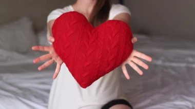 Örgü kırmızı kalp taşıyan kız, yakın plan. Sevgililer Günü. Kalp şeklinde yastık. Sevgililer günü hediyesi. Oyuncak kalple yatakta oturan bir kız. Aşkın sembolü, hediye. Yastıklı romantik genç kadın