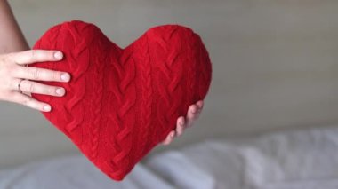 Örgü kırmızı kalp taşıyan kız, yakın plan. Sevgililer Günü. Kalp şeklinde yastık. Sevgililer günü hediyesi. Oyuncak kalple yatakta oturan bir kız. Aşkın sembolü, hediye. Yastıklı romantik genç kadın