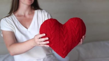 Örgü kırmızı kalp taşıyan kız, yakın plan. Sevgililer Günü. Kalp şeklinde yastık. Sevgililer günü hediyesi. Oyuncak kalple yatakta oturan bir kız. Aşkın sembolü, hediye. Yastıklı romantik genç kadın