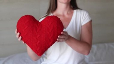 Örgü kırmızı kalp taşıyan kız, yakın plan. Sevgililer Günü. Kalp şeklinde yastık. Sevgililer günü hediyesi. Oyuncak kalple yatakta oturan bir kız. Aşkın sembolü, hediye. Yastıklı romantik genç kadın