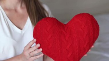 Örgü kırmızı kalp taşıyan kız, yakın plan. Sevgililer Günü. Kalp şeklinde yastık. Sevgililer günü hediyesi. Oyuncak kalple yatakta oturan bir kız. Aşkın sembolü, hediye. Yastıklı romantik genç kadın