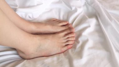 Beyaz çarşafların üzerinde kadın bacakları var. Dişi ayakları. Battaniyenin altından bakan çıplak ayaklı bir kız. Bacaklar beyaz çarşafların üzerinde parmaklarını oynatıyor. Dinlen.