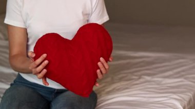 Örgü kırmızı kalp taşıyan kız, yakın plan. Sevgililer Günü. Kalp şeklinde yastık. Sevgililer günü hediyesi. Oyuncak kalple yatakta oturan bir kız. Aşkın sembolü, hediye. Yastıklı romantik genç kadın