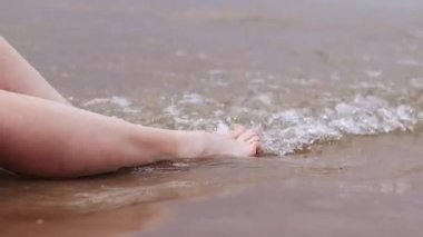 Sahilde kadın bacakları, deniz, yakın plan. Deniz dalgalarının arka planında çıplak ayaklar. Kadın bacakları yakın çekim, plaj tatili. Kumsalda dinlenmek, yaz tatili.
