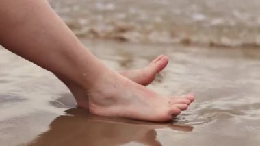 Sahilde kadın bacakları, deniz, yakın plan. Deniz dalgalarının arka planında çıplak ayaklar. Kadın bacakları yakın çekim, plaj tatili. Kumsalda dinlenmek, yaz tatili.