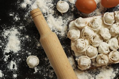 Çiğ pelmeni, üst görüş, düz yatış. Pelmeni, un, yumurta ve tahta raptiye. Tahta tahtada etli börek ya da ravioli, karanlık masa. Geleneksel Slav yemeği. Gıda arkaplanı