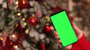 Kadın elinde yeşil ekran telefon. Noel ağacının arka planında. Bir kadın elinin yakın çekimi ve yeşil ekranlı bir akıllı telefon, yeni yıl. Modern cep telefonu. Modelleme. Teknoloji
