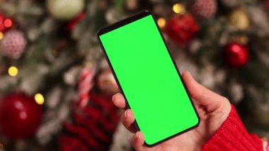 Kadın elinde yeşil ekran telefon. Noel ağacının arka planında. Bir kadın elinin yakın çekimi ve yeşil ekranlı bir akıllı telefon, yeni yıl. Modern cep telefonu. Modelleme. Teknoloji
