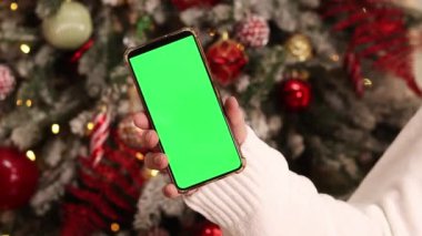 Kadın elinde yeşil ekran telefon. Noel ağacının arka planında. Bir kadın elinin yakın çekimi ve yeşil ekranlı bir akıllı telefon, yeni yıl. Modern cep telefonu. Modelleme. Teknoloji