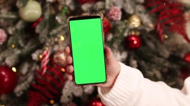 Kadın elinde yeşil ekran telefon. Noel ağacının arka planında. Bir kadın elinin yakın çekimi ve yeşil ekranlı bir akıllı telefon, yeni yıl. Modern cep telefonu. Modelleme. Teknoloji
