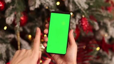 Kadın elinde yeşil ekran telefon. Noel ağacının arka planında. Bir kadın elinin yakın çekimi ve yeşil ekranlı bir akıllı telefon, yeni yıl. Modern cep telefonu. Modelleme. Teknoloji