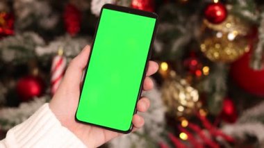 Kadın elinde yeşil ekran telefon. Noel ağacının arka planında. Bir kadın elinin yakın çekimi ve yeşil ekranlı bir akıllı telefon, yeni yıl. Modern cep telefonu. Modelleme. Teknoloji