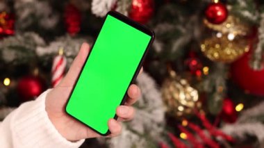 Kadın elinde yeşil ekran telefon. Noel ağacının arka planında. Bir kadın elinin yakın çekimi ve yeşil ekranlı bir akıllı telefon, yeni yıl. Modern cep telefonu. Modelleme. Teknoloji