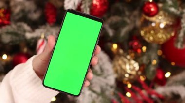 Kadın elinde yeşil ekran telefon. Noel ağacının arka planında. Bir kadın elinin yakın çekimi ve yeşil ekranlı bir akıllı telefon, yeni yıl. Modern cep telefonu. Modelleme. Teknoloji