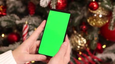 Kadın elinde yeşil ekran telefon. Noel ağacının arka planında. Bir kadın elinin yakın çekimi ve yeşil ekranlı bir akıllı telefon, yeni yıl. Modern cep telefonu. Modelleme. Teknoloji