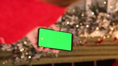 Kadın elinde yeşil ekran telefon. Noel ağacının arka planında. Bir kadın elinin yakın çekimi ve yeşil ekranlı bir akıllı telefon, yeni yıl. Modern cep telefonu. Modelleme. Teknoloji