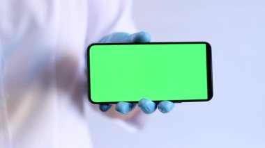 Mavi eldivenli bir kadın. Elinde yeşil ekranlı bir telefon var. Elinde akıllı telefon olan doktor ya da hemşire. Mavi eldivenli ve beyaz ceketli doktor cep telefonu tutuyor. Model