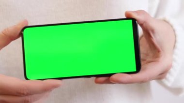 Yeşil ekranlı cep telefonu. Yeşil ekranlı akıllı telefonu tutan kadın eller, yakın plan. Beyaz arka planda telefonu olan beyaz kazaklı bir kız. Kromakey cep telefonunu tutan tanımlanamayan el