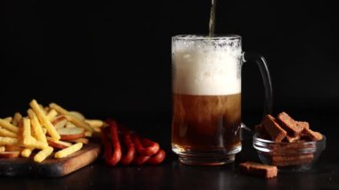 Hafif bira ve koyu arkaplanda atıştırmalıklar. Bira, patates kızartması ve av sosisi. Tahtada cam ve aperatifler. Hafif bira ve kızarmış ekmek. Geleneksel cam bira kupası.