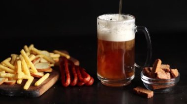 Hafif bira ve koyu arkaplanda atıştırmalıklar. Bira, patates kızartması ve av sosisi. Tahtada cam ve aperatifler. Hafif bira ve kızarmış ekmek. Geleneksel cam bira kupası.