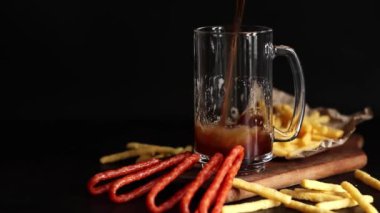 Koyu renkli bira ve arka planda atıştırmalıklar. Bira, patates kızartması ve av sosisi. Ahşap tahta üzerinde cam ve aperatifler, yakın plan. Koyu bira ve kızarmış ekmek. Geleneksel cam bira kupası.