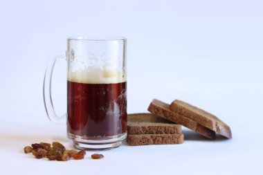 Bir kupa lezzetli kvass, hafif arka planda ekmek ve kuru üzüm. Cam bardak kvass köpüklü, yakın plan. Beyaz arka planda malzemeleri olan Kvass. Ekmek dilimleriyle taze içecek, seçici odaklanma.