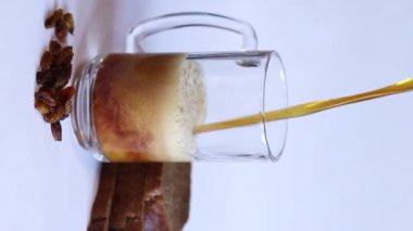 Bir kupa lezzetli kvass, hafif arka planda ekmek ve kuru üzüm. Cam bardak kvass köpüklü, yakın plan. Beyaz arka planda malzemeleri olan Kvass. Ekmek dilimleriyle taze içecek, seçici odaklanma.