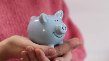 Pembe, sıcak kazaklı bir kadın kumbarayı elinde tutuyor, yakın plan. Domuz kumbarası kadınların elinde. Mavi domuz kumbarası, tasarruf ve finansal planlama kavramı. Şirin domuz kumbarasının yakın çekimi.