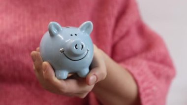 Pembe, sıcak kazaklı bir kadın kumbarayı elinde tutuyor, yakın plan. Domuz kumbarası kadınların elinde. Mavi domuz kumbarası, tasarruf ve finansal planlama kavramı. Şirin domuz kumbarasının yakın çekimi.