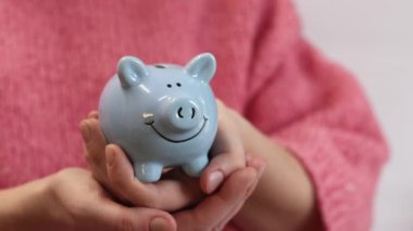 Pembe, sıcak kazaklı bir kadın kumbarayı elinde tutuyor, yakın plan. Domuz kumbarası kadınların elinde. Mavi domuz kumbarası, tasarruf ve finansal planlama kavramı. Şirin domuz kumbarasının yakın çekimi.
