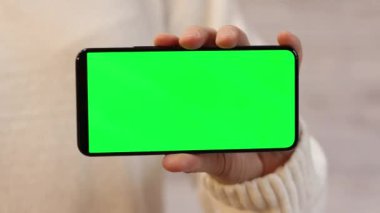 Elinde yeşil ekranlı bir telefon var. Elinde akıllı telefon tutan beyaz kazaklı bir kadın. Modern aletler. Cep telefonunun yakın çekimi. Krom anahtar, internette sörf yapmak, içeriği görmek