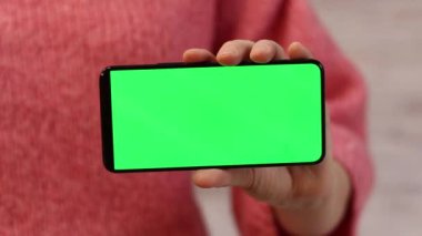 Elinde yeşil ekranlı bir telefon var. Elinde akıllı telefon tutan pembe kazaklı bir kadın. Modern aletler. Cep telefonunun yakın çekimi. Krom anahtar, internette sörf yapmak.