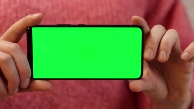 Elinde yeşil ekranlı bir telefon var. Elinde akıllı telefon tutan pembe kazaklı bir kadın. Modern aletler. Cep telefonunun yakın çekimi. Krom anahtar, internette sörf yapmak.